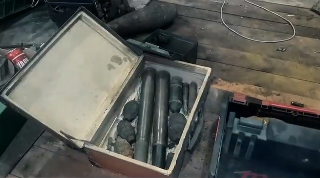 Skrzynka z metalowymi przedmiotami, w tym pociskami i granatami, leży na drewnianej podłodze w warsztacie.