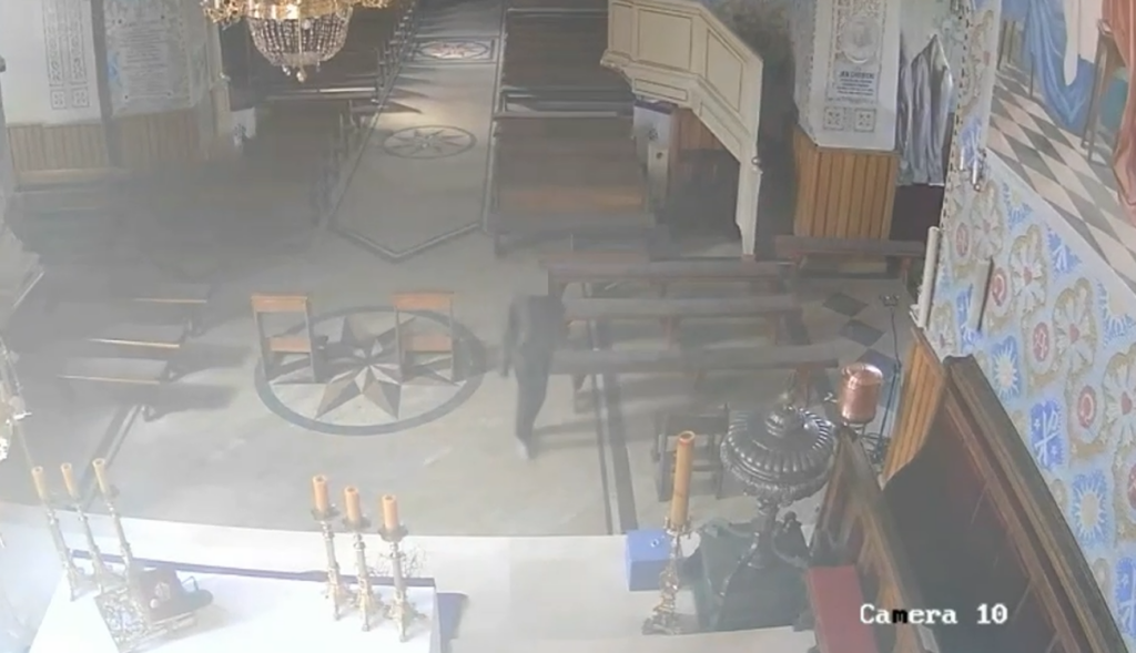 Mężczyzna w ciemnym ubraniu przechodzi przez wnętrze kościoła, mijając ławki i świeczniki.