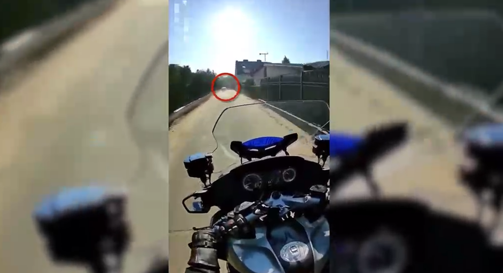Mężczyzna na motocyklu jedzie wzdłuż pustej drogi, w tle widoczny jest dom i ogrodzenie.