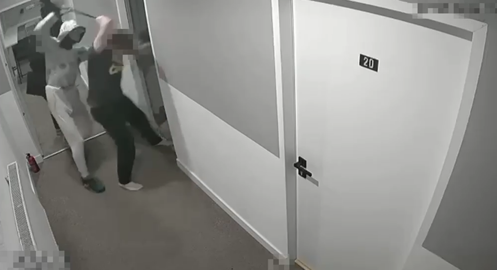 Osoba w białym kapturze atakuje drugą osobę w korytarzu, przy drzwiach oznaczonych numerem 20.