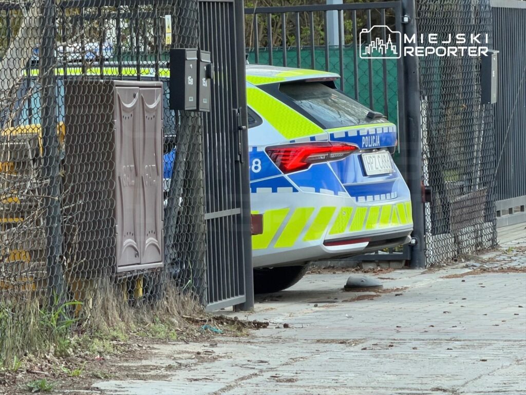 Policyjny samochód zaparkowany za ogrodzeniem w pobliżu zamkniętej bramy.