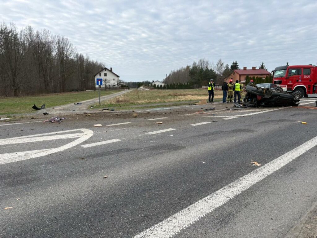 Na drodze leżą resztki wozu po wypadku, a strażacy oraz policjanci prowadzą działania ratunkowe. W tle widoczny dom.