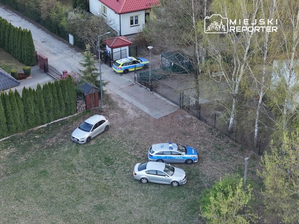 Na placu przed domem parkują dwa samochody osobowe oraz radiowóz policyjny, w tle widać ogrodzenie i drzewa.