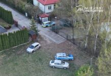 Na placu przed domem parkują dwa samochody osobowe oraz radiowóz policyjny, w tle widać ogrodzenie i drzewa.