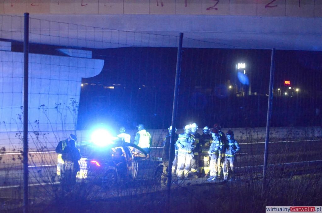 Grupa strażaków i ratowników medycznych przy samochodzie osobowym na drodze, oświetlona niebieskimi światłami.