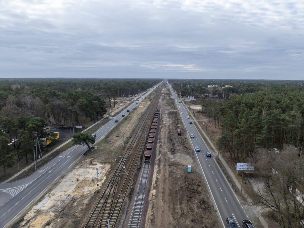 Widok na torowisko kolejowe z pociągami towarowymi, obok ruchliwa droga i las w tle.