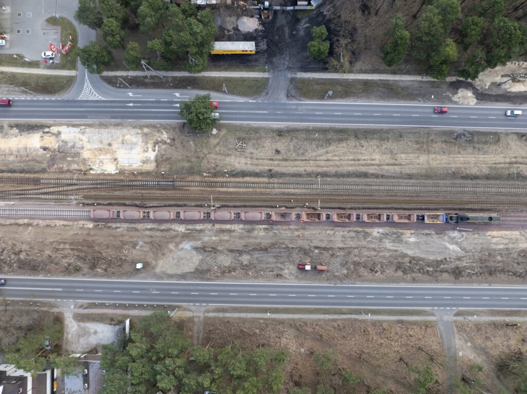 Pojazdy ciężarowe poruszają się wzdłuż drogi obok torów kolejowych, w otoczeniu wycinki drzew.