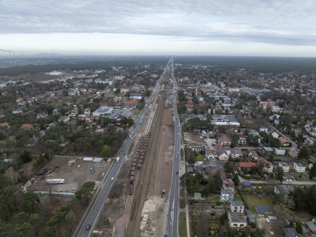 Widok na tory kolejowe z wagonami towarowymi, obok droga z samochodami i zabudowa mieszkalna w tle.