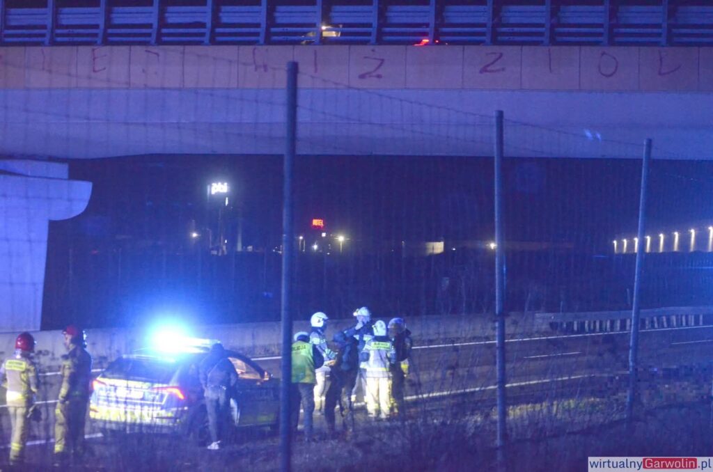 Grupa ratowników i policjantów prowadzi działania na drodze w nocy, obok zaparkowanego radiowozu z włączonymi sygnalizatorami.