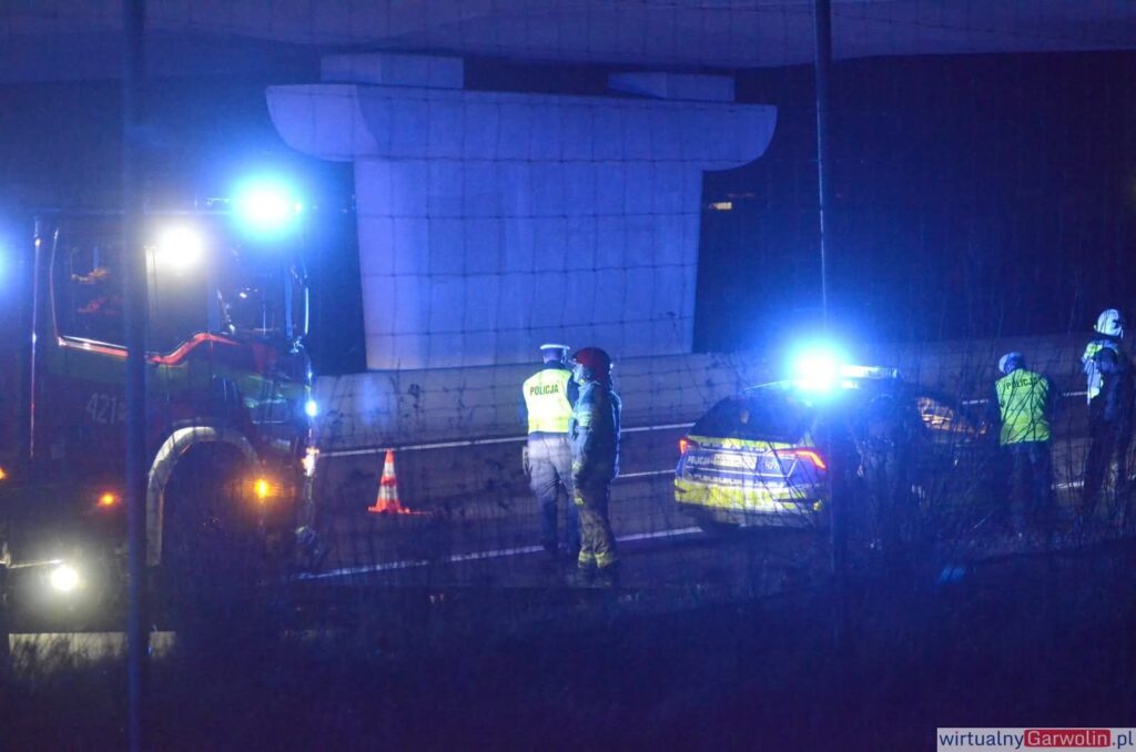 Policjant rozmawia z ratownikiem na oświetlonej drodze, obok stoi wóz strażacki i radiowóz z włączonymi sygnałami.