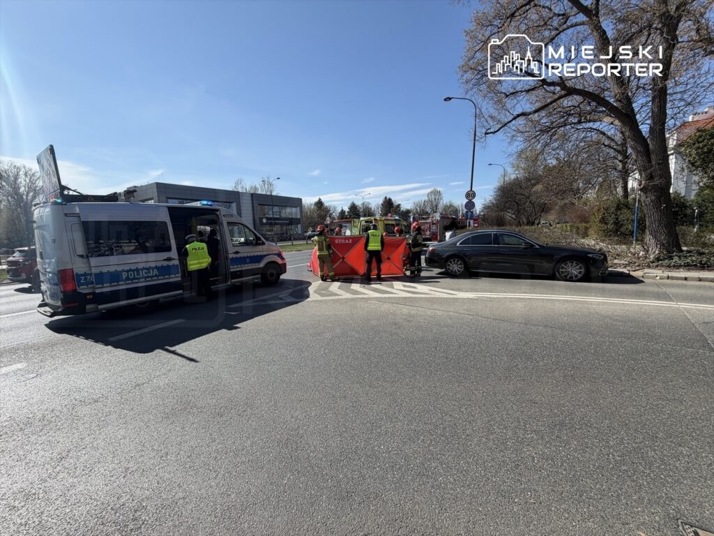 Policjant rozmawia z kierowcą przy oznakowanym samochodzie policyjnym, obok blokady drogi i strażaków w kamizelkach.