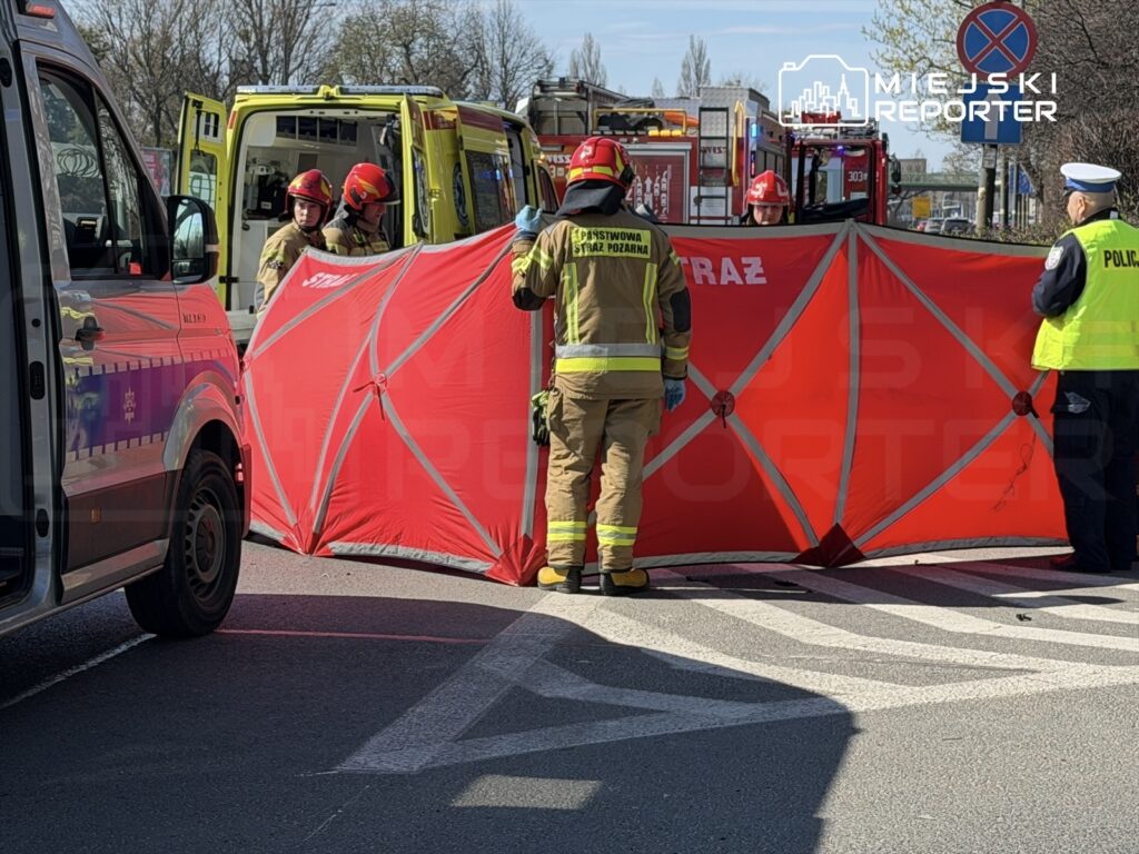 Grupa strażaków w hełmach ustawia czerwoną osłonę na jezdni, obok pojazdów ratunkowych i policji.