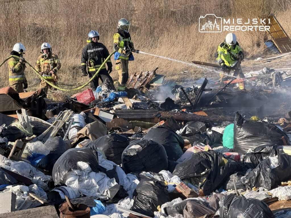 Czterech strażaków w hełmach gasi pożar na wysypisku, otoczonym stosami odpadków i dymem.