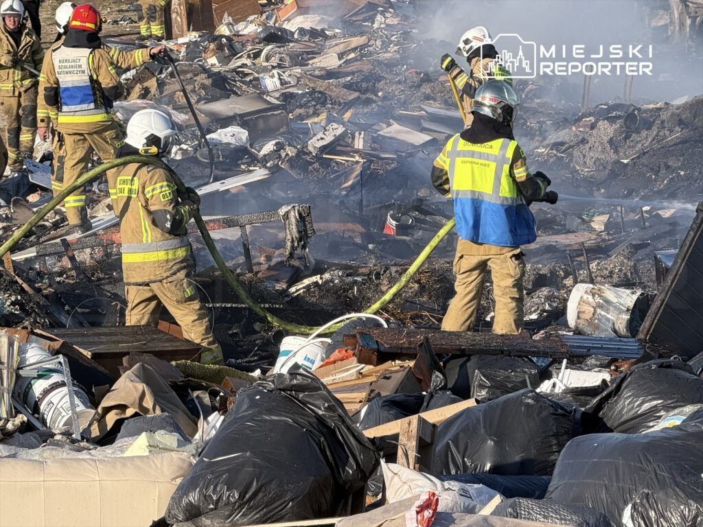 Strażacy gaszą pożar w zniszczonym obszarze, przeszukując zgliszcza wśród odpadów i dymu.