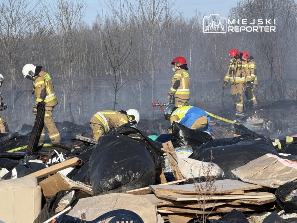 Grupa strażaków w mundurach gaśniczych przeszukuje teren z odpadami, gdzie unosi się dym.