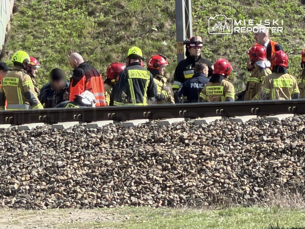 Grupa ratowników i policjantów udziela pomocy osobie na torach kolejowych w otoczeniu kamieni.