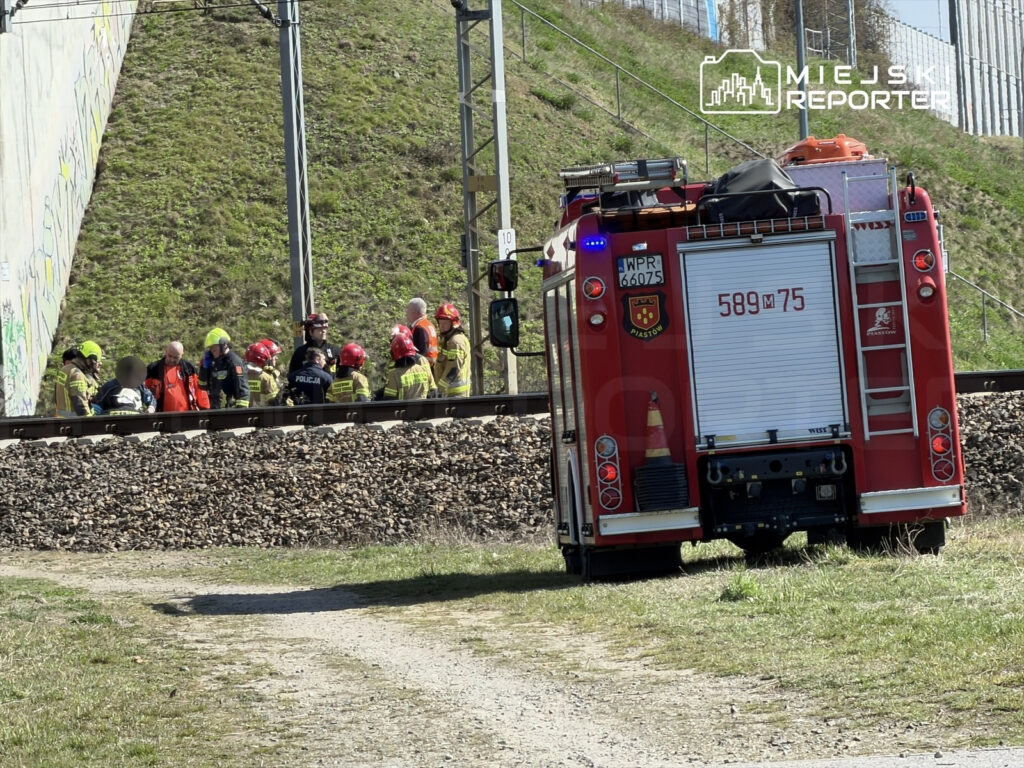 Grupa ratowników w kaskach omawia sytuację obok torów kolejowych, w tle widoczny wóz strażacki.