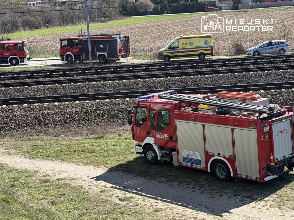Dwa wozy strażackie i karetka stoją obok torów kolejowych na zielonym terenie.