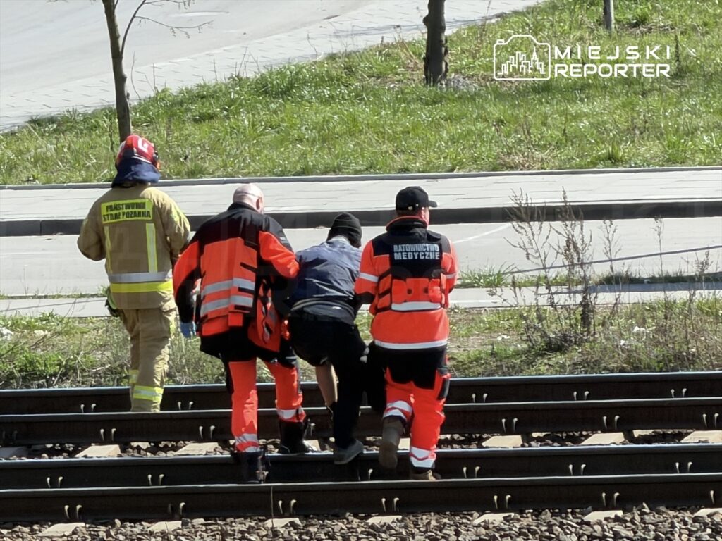 Grupa ratowników medycznych i strażaków pomaga osobie poszkodowanej przy torach kolejowych, w słoneczny dzień.