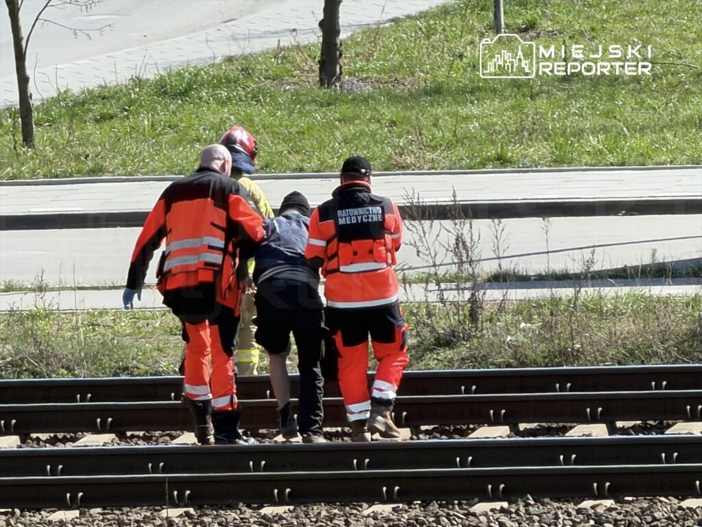 Czterech ratowników medycznych pomaga osobie w trudnej sytuacji na torach kolejowych.