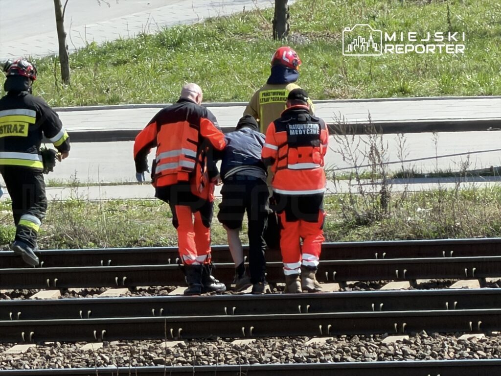 Ratownicy medyczni i strażacy pomagają osobie na torach kolejowych w słoneczny dzień.
