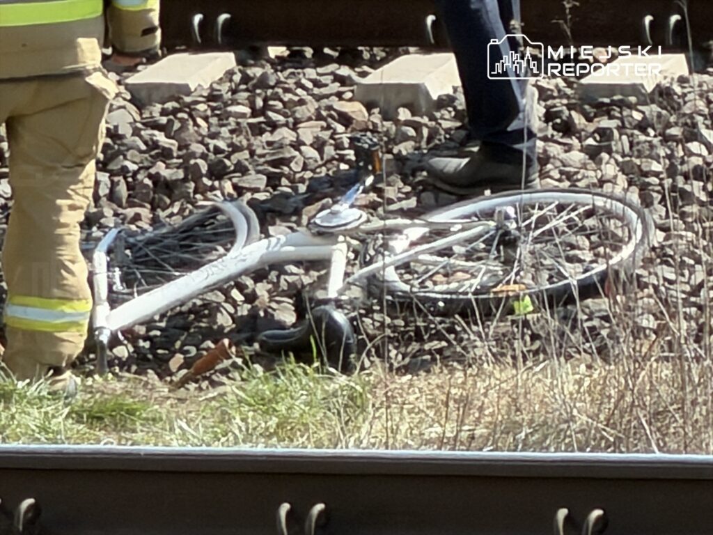 Strażak w mundurze stoi obok leżącego roweru na torach kolejowych, w otoczeniu kamieni i trawy.