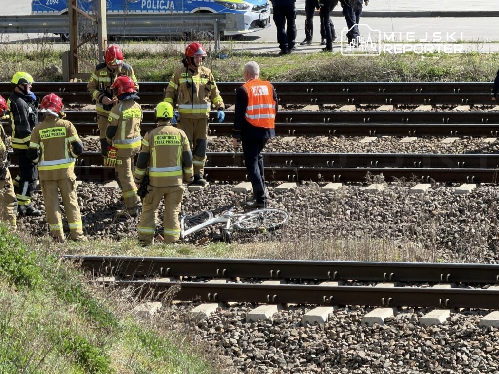 Grupa strażaków w mundurach z odblaskowymi elementami zbiera się przy torach kolejowych, obok leżącego roweru.