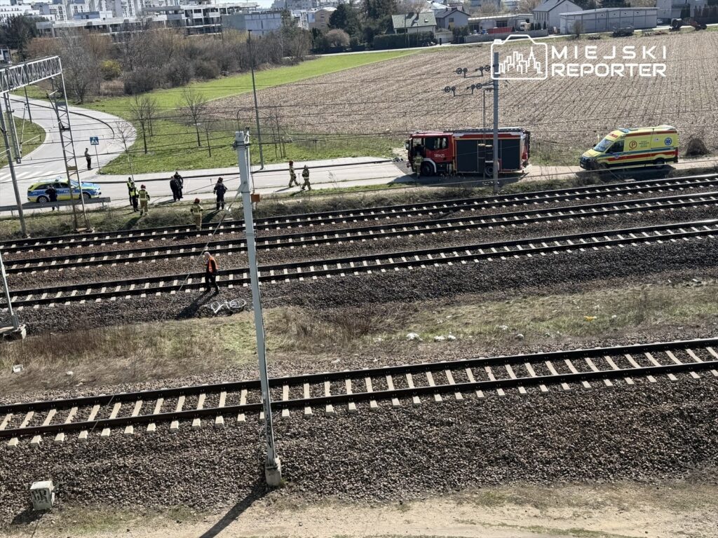 Grupa ratowników i policjantów pracuje przy torach kolejowych, obok zaparkowanej straży pożarnej i karetki.