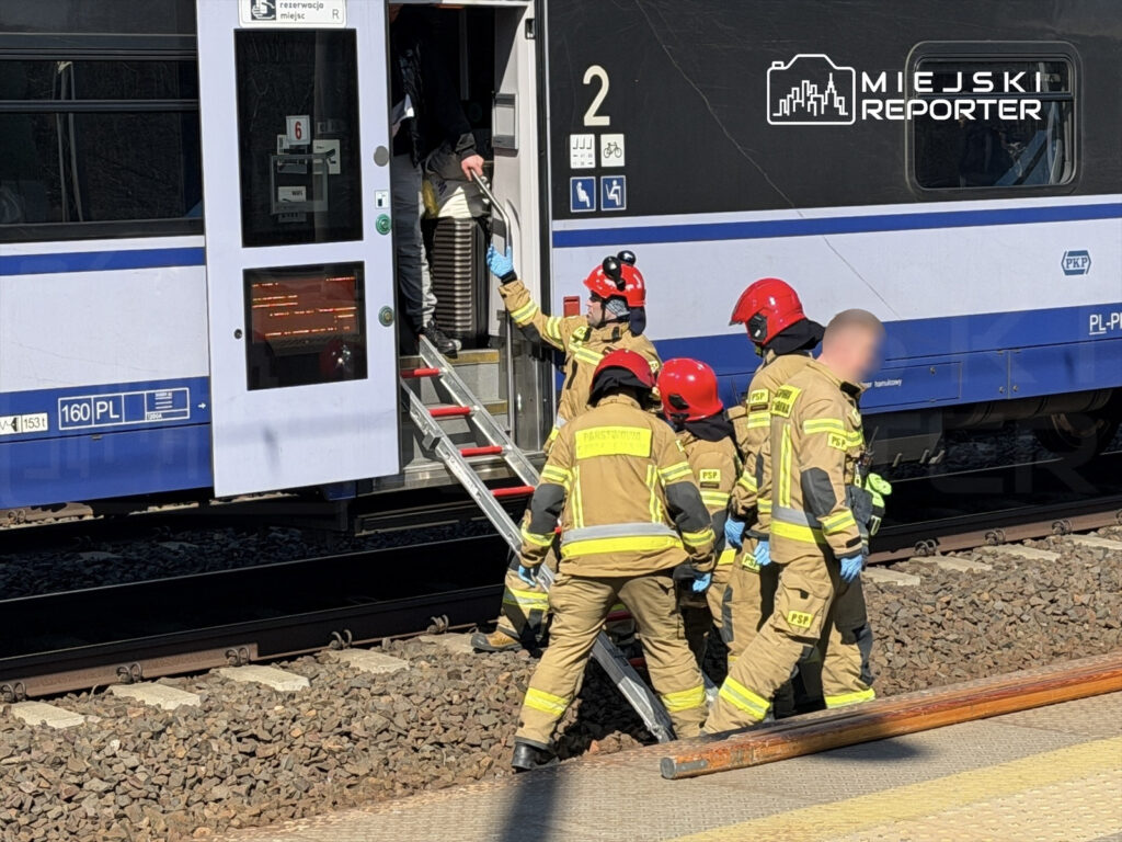 Grupa strażaków w uniformach z hełmami na głowach pomaga osobie opuszczającej pociąg na stacji kolejowej.