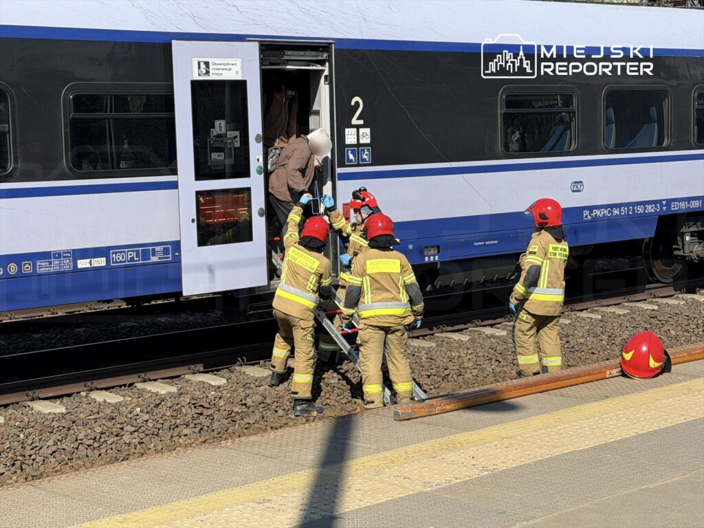 Grupa strażaków w kaskach pomaga osobie opuszczającej pociąg na stacji kolejowej.