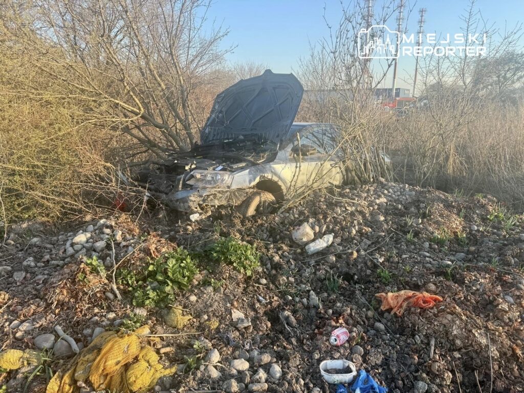 Zniszczony samochód z otwartą maską, zaparkowany w zaroślach obok wysypiska śmieci.