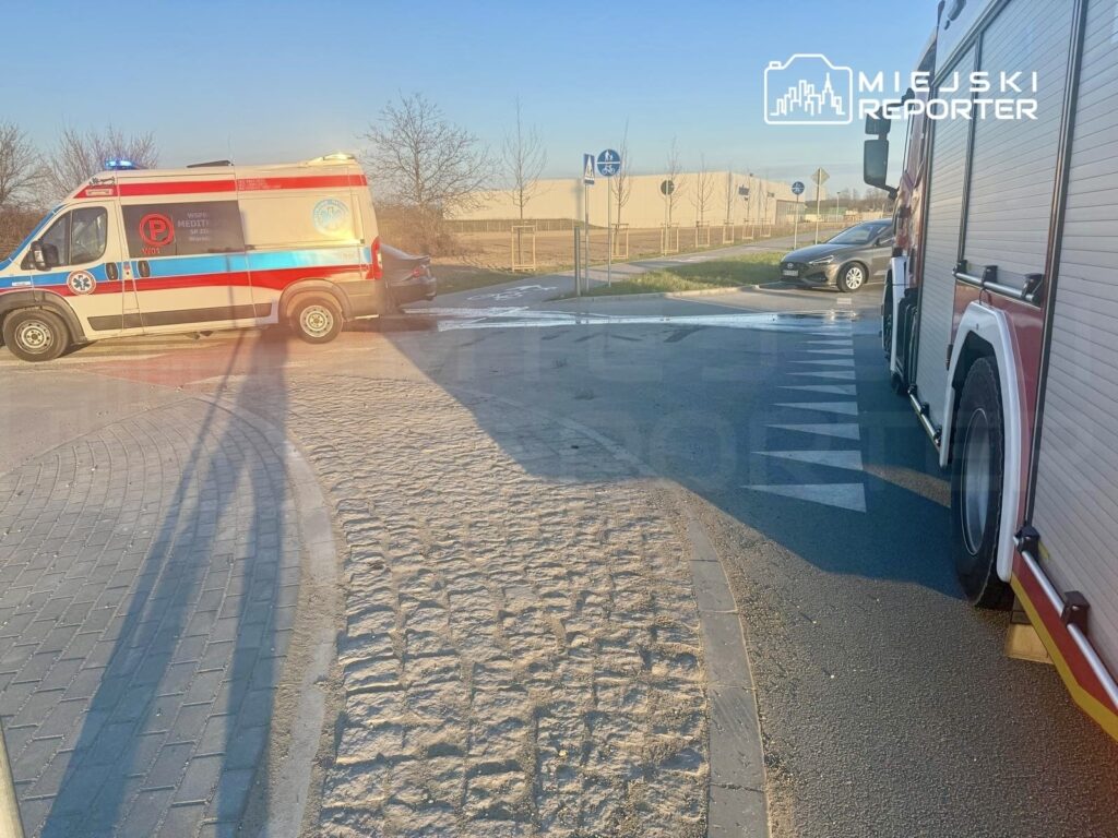 Karetka pogotowia oraz wóz strażacki na drodze, w tle widać budynki i znaki drogowe.