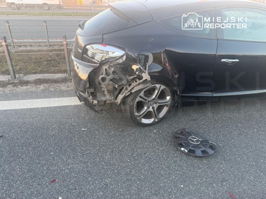 Czarny samochód osobowy z uszkodzoną tylną częścią stoi na poboczu drogi, obok leży odpadnięta osłona koła.