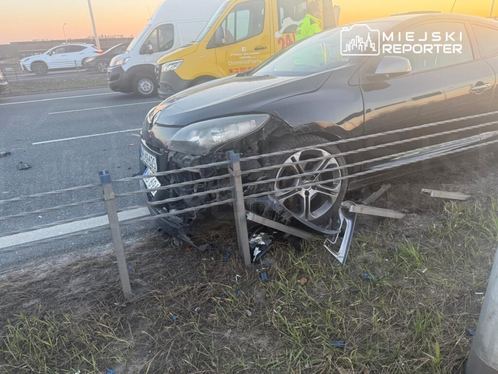 Czarny samochód osobowy wjeżdżający w barierki ochronne na poboczu drogi, obok ruchliwej jezdni.