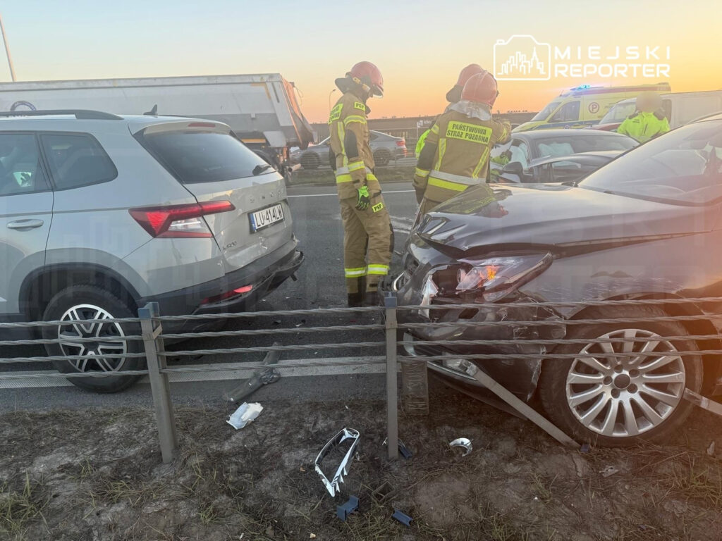 Dwaj strażacy w żółtych mundurach badają skutki wypadku drogowego na autostradzie, obok uszkodzonych samochodów.