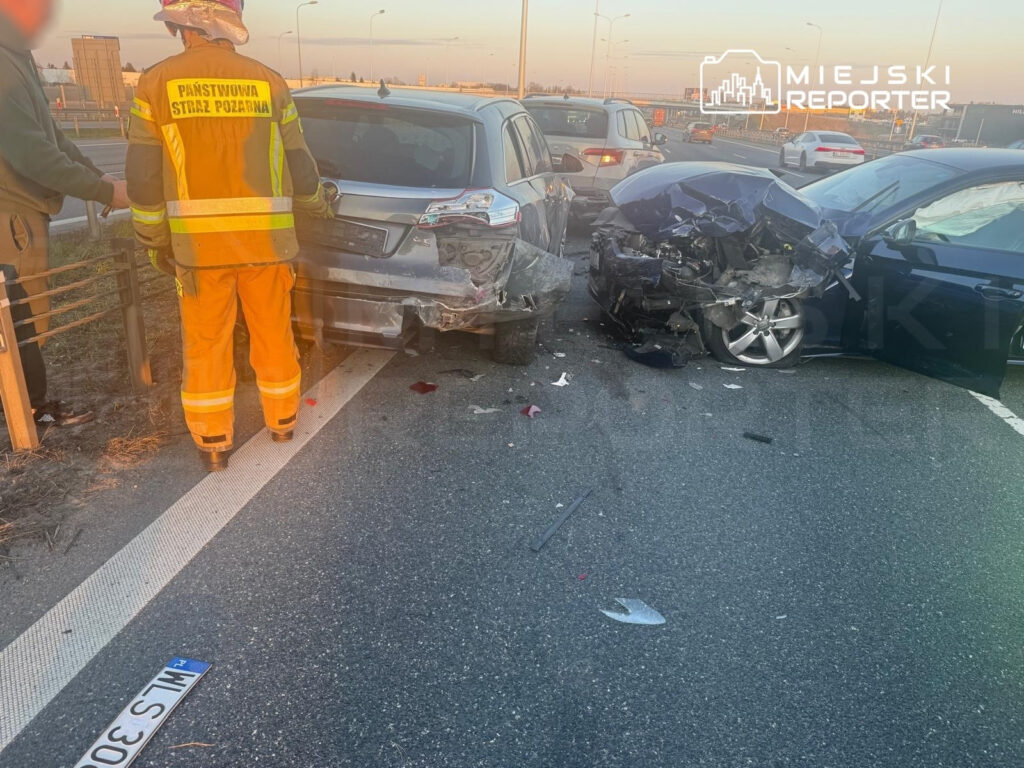 Strażak w pomarańczowym mundurze ocenia skutki wypadku drogowego, w którym zderzyły się dwa samochody na jezdni.