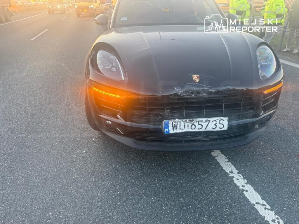 Czarny samochód marki Porsche z uszkodzoną przednią częścią stoi na drodze, w tle widoczne inne pojazdy oraz osoby w odblaskowych kamizelkach.