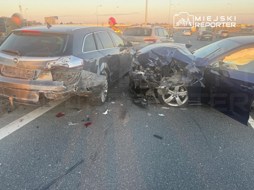 Dwa uszkodzone samochody osobowe zderzone na pasie drogi, obok stoją strażacy w mundurach.