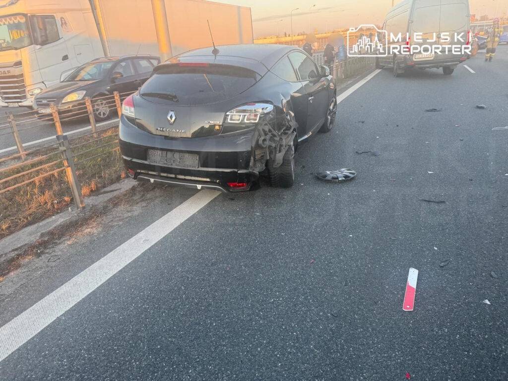 Czarny samochód osobowy z uszkodzeniami stoi na poboczu drogi, obok leżą fragmenty opon i innych detali.