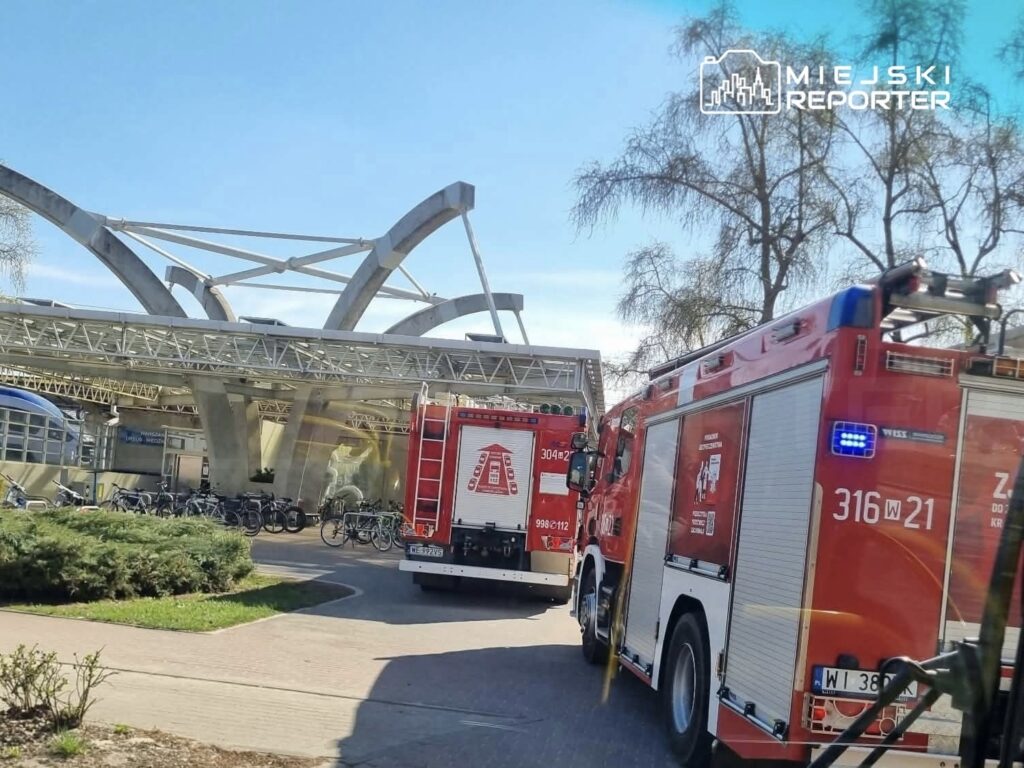 Wozy strażackie z włączonymi sygnałami dojeżdżają do nowoczesnego budynku z przeszkloną fasadą.
