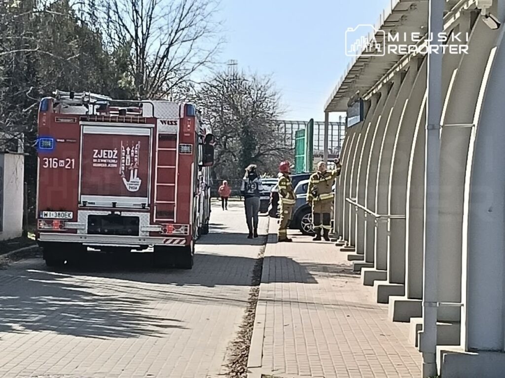 Czerwony wóz strażacki stoi przy chodniku, a strażacy w mundurach rozmawiają obok budynku.
