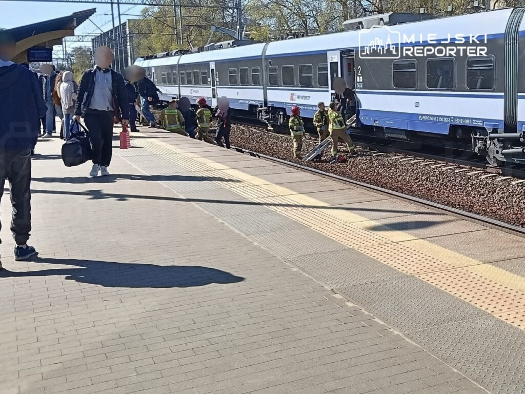 Grupa strażaków pracuje przy torach kolejowych, obok pociągu, na stacji w słoneczny dzień. Pasażerowie czekają na peronie.