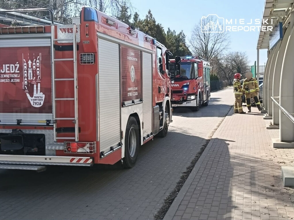 Czerwony wóz strażacki zaparkowany przy chodniku, obok dwóch strażaków rozmawiających w mundurach ochronnych.