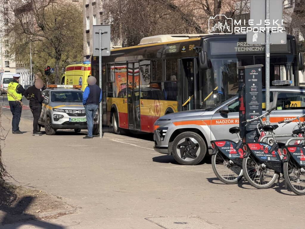 Policjant rozmawia z mężczyzną obok samochodu służbowego, obok zatrzymany autobus i stacja rowerów miejskich.
