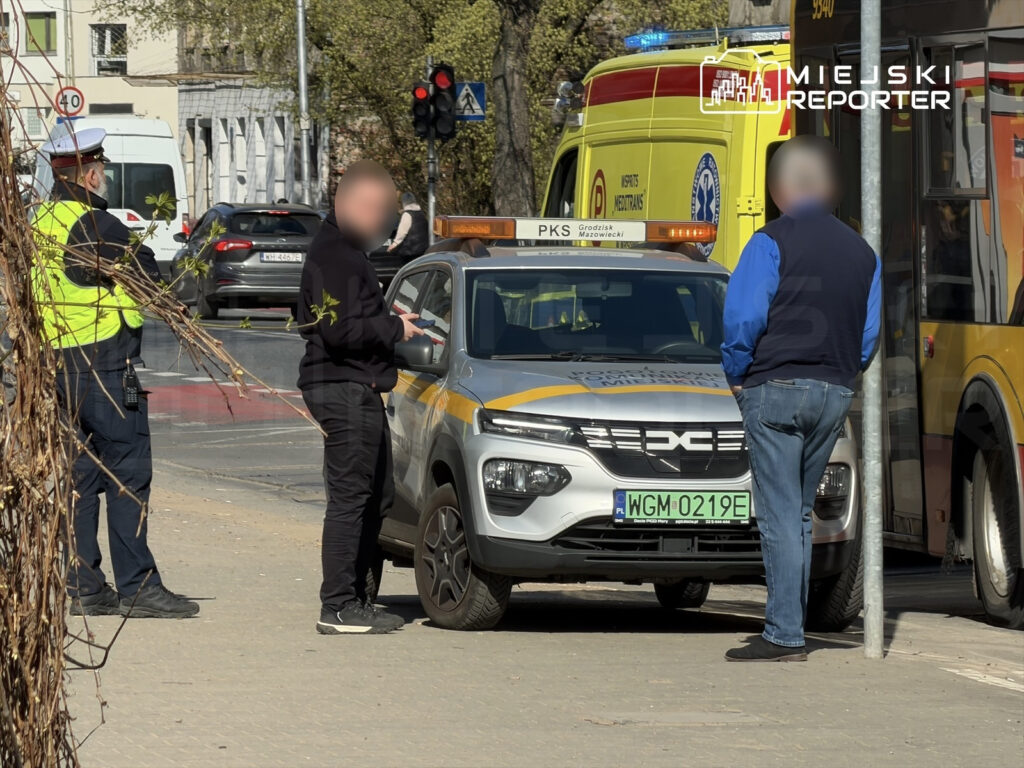 Policjant w kamizelce odblaskowej rozmawia z mężczyzną przy oznakowanym samochodzie służbowym, obok przejeżdżających pojazdów.