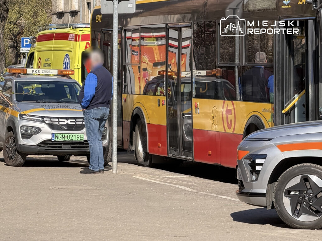 Mężczyzna w niebieskiej koszuli rozmawia obok taksówki z oznaczeniem, w tle autobus i pojazdy służb ratunkowych.
