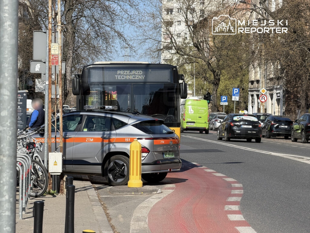Autobus z napisem "PRZEJAZD TECHNICZNY" zatrzymany obok policyjnego pojazdu na ulicy, w tle widać zaparkowane samochody.