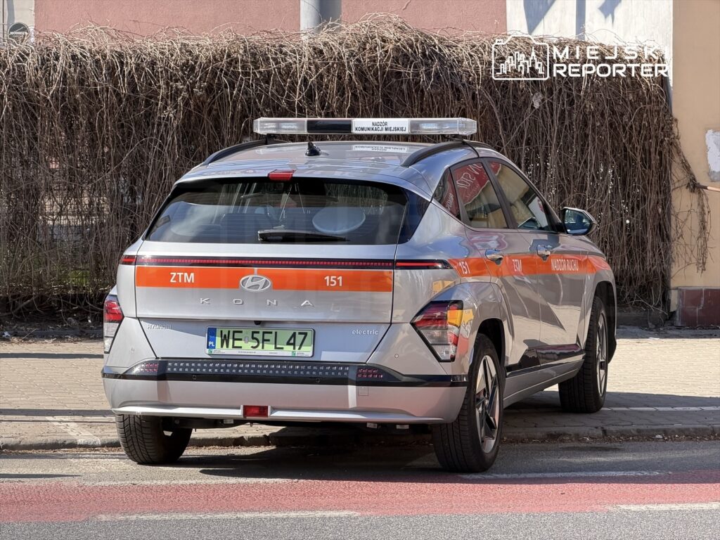 Elektryczny samochód służbowy ZTM z napisem "Nadzór Ruchu" zaparkowany na chodniku w miejskim otoczeniu.