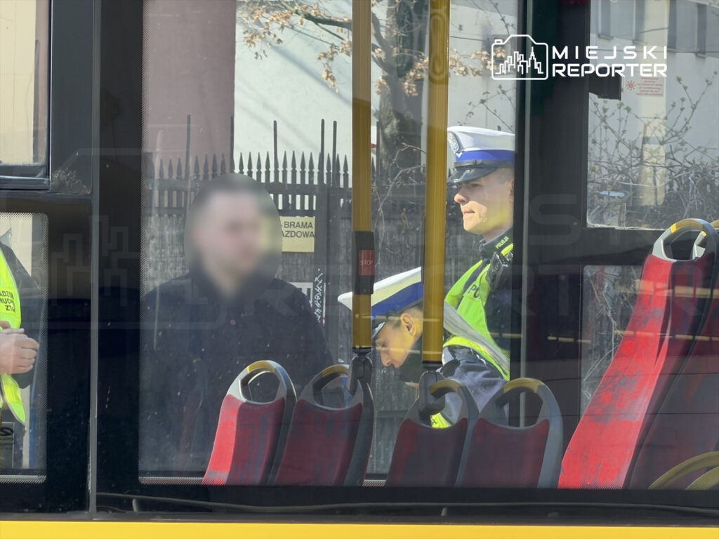 Mężczyzna w czarnej odzieży stoi obok dwóch policjantów w mundurach, wewnątrz autobusu miejskiego.