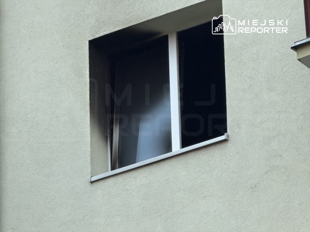 Okno w ścianie budynku z zasłoniętą roletą, widoczne na tle jasnej elewacji.